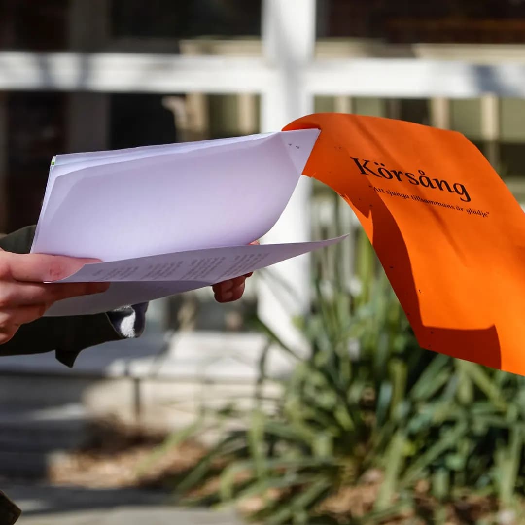 Händer håller noter framför en orange vimpel med texten Körsång