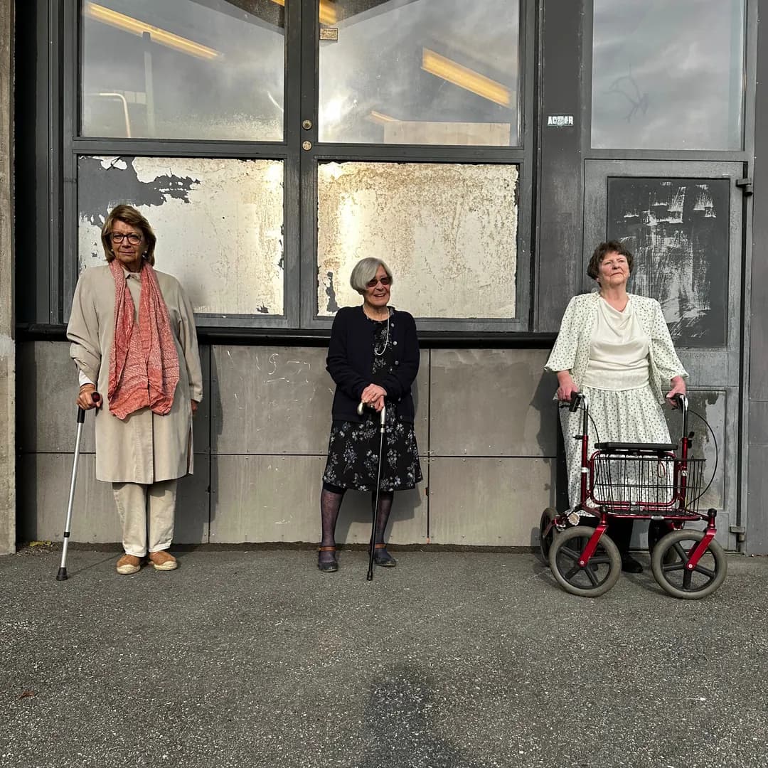 Tre äldre kvinnor med käpp och rollator står i solen framför en husvägg