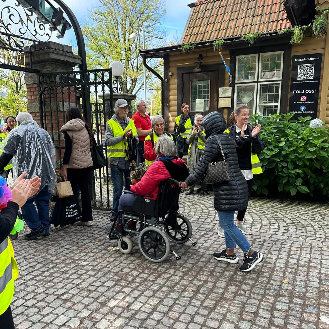 Äldre och personal i gula västar på utflykt vid en entré med smidesgrindar