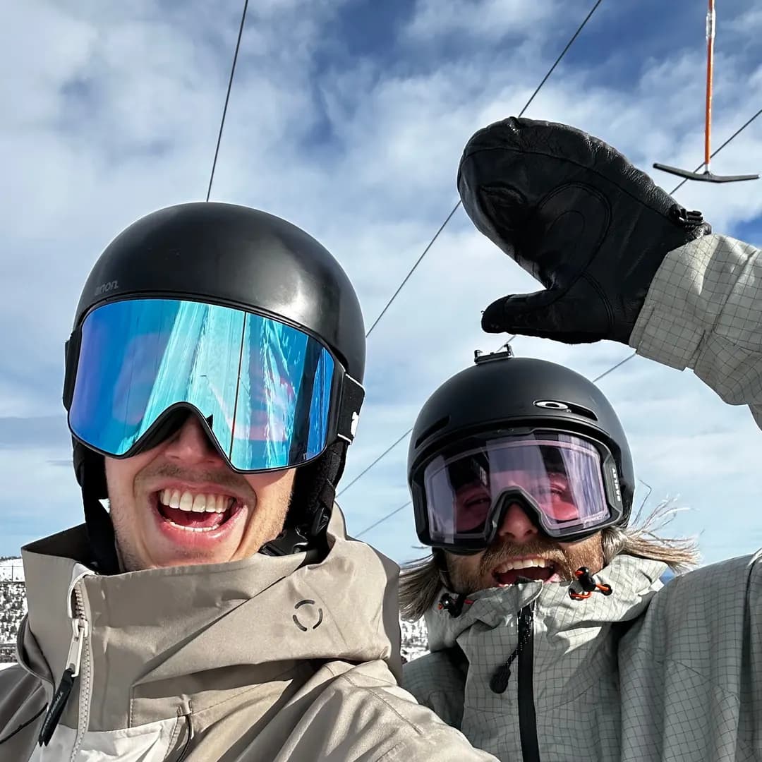 Två skidåkare med hjälmar och goggles tar en glad selfie i liften